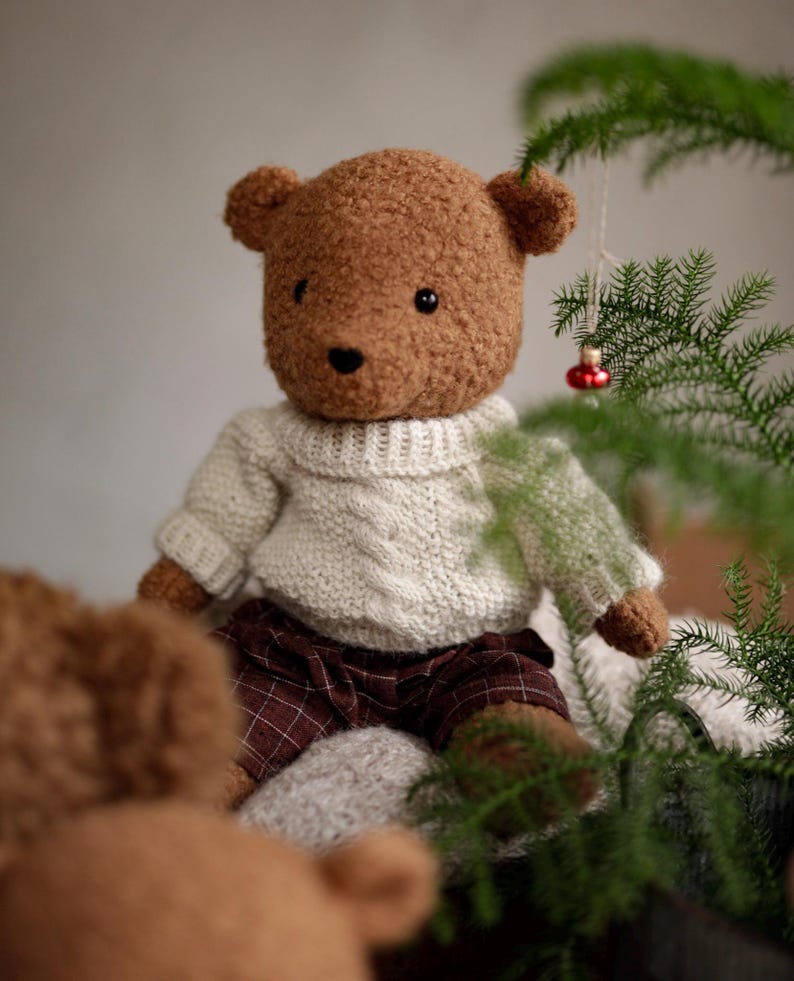 May include: A brown teddy bear wearing a white knitted sweater and plaid pants sits near a green plant with a red ornament.