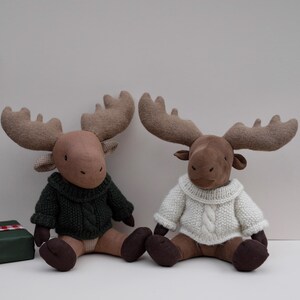 May include: Two brown plush moose wearing knitted sweaters. One moose is wearing a green sweater and the other is wearing a white sweater.