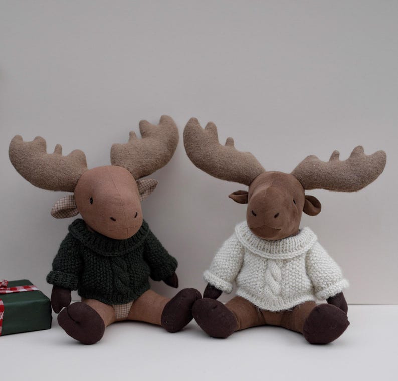 May include: Two brown plush moose wearing knitted sweaters. One moose is wearing a green sweater and the other is wearing a white sweater.