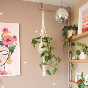 Pode incluir: Uma parede rosa com um suporte de plantas de macramê com uma planta verde pendurada nele. Há várias plantas em vasos sobre uma mesa branca em frente à parede. Há uma impressão emoldurada com uma cena de pôr do sol sobre a mesa. Há uma bola de discoteca pendurada no teto.