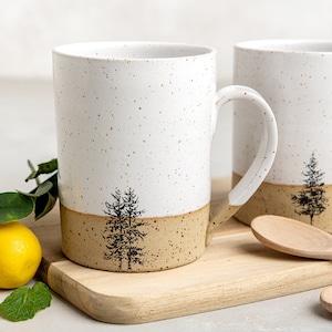 May include: Two ceramic mugs featuring a white speckled upper section and a tan speckled lower section. Each mug displays a black ink illustration of a pine tree. The mugs have a cylindrical shape with a curved handle on the side for gripping.