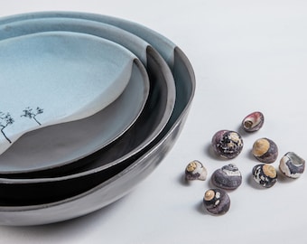 May include: A set of three nested ceramic bowls with a two-tone finish, featuring a light blue interior with a black tree silhouette and a dark charcoal exterior. Beside the bowls are seven small, dark, textured sea snail shells.