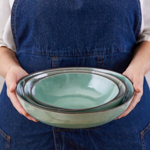 May include: A set of two nested ceramic bowls with a smooth, light seafoam green glaze and a dark, contrasting rim. The bowls feature a rounded, organic shape with a glossy finish, stacked one inside the other to display their varying sizes.