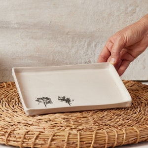 Peut inclure: Un plateau en céramique blanche avec un motif simple de deux arbres imprimés dessus. Le plateau est posé sur un dessous de plat en paille tressée.