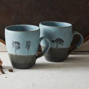 May include: Two blue ceramic mugs with black tree silhouettes and a dark grey band around the bottom. The mugs are on a white surface with coffee beans and a stack of chocolate and pink layered squares.