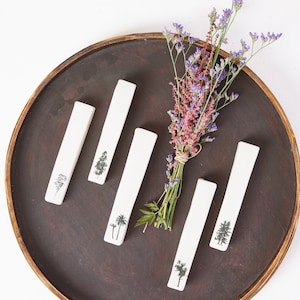 Könnte beinhalten: Fünf weiße Keramik-Lesezeichen mit verschiedenen Baumdesigns auf einem braunen Holztablett. Ein Strauß lila Blumen befindet sich in der Mitte des Tabletts.