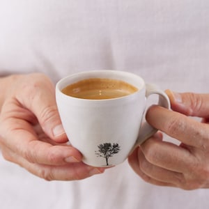 以下が含まれることがあります： 表面に質感のあるマットなオフホワイトの陶器製コーヒーカップで、前面に小さな黒い木のシルエットがプリントされています。カップには表面に薄茶色のクレマ層があるダークブラウンのコーヒーが入っており、持ち手が付いています。
