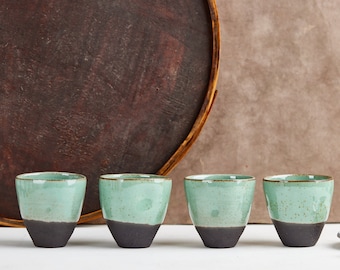 May include: Four ceramic cups arranged in a row, each featuring a smooth, light mint green glazed upper section and a contrasting matte dark charcoal base. The cups have a tapered, handleless design with a subtle speckled texture on the glaze.