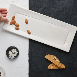 May include: A white rectangular serving platter with a slightly raised edge, holding a small pile of almonds. The platter is on a white surface with a black surface to the right.