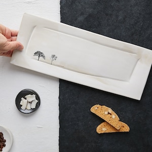 Peut inclure: Un plat de service rectangulaire en céramique blanche avec un bord surélevé et une surface texturée subtile. Le plat présente une silhouette noire de deux arbres avec le texte "Little Forest" imprimé en dessous.