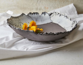 May include: A shallow, wide ceramic bowl with a dark gray exterior and a white interior featuring black crackle patterns. The rim is uneven and textured, resembling natural stone. Four bright yellow marigold flowers rest inside the bowl.