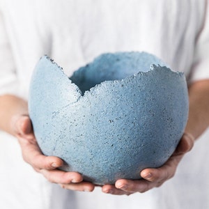 May include: A blue, textured, spherical bowl with a jagged rim. The bowl is held in both hands against a white background. The bowl's colour is a muted, dusty blue.
