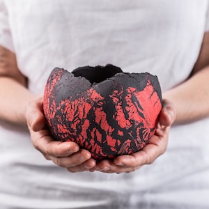 May include: A round, bowl-shaped ceramic vessel with a rough, uneven rim. The exterior features a textured, matte black surface with deep, jagged cracks that reveal a vibrant, bright red color underneath, creating a high-contrast, volcanic appearance.