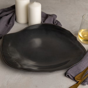 May include: A large, matte black ceramic dinner plate with an irregular, organic, wavy rim shape. The plate features a smooth, dark surface with subtle light reflections, showcasing a handcrafted aesthetic that contrasts with the dark gray fabric.