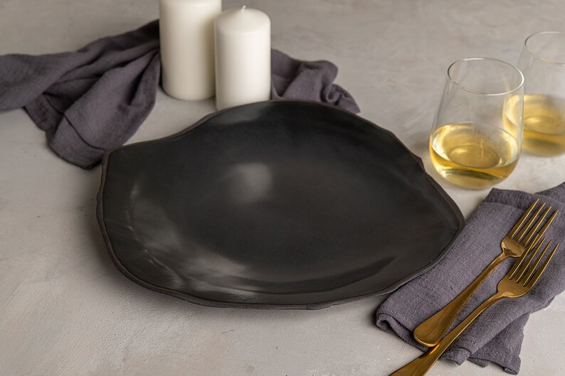 May include: A large, matte black ceramic dinner plate with an irregular, organic, wavy rim shape. The plate features a smooth, dark surface with subtle light reflections, showcasing a handcrafted aesthetic that contrasts with the dark gray fabric.