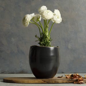 May include: White ranunculus flowers in a black ceramic vase on a wooden surface.