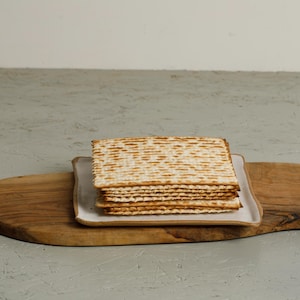 May include: A stack of matzah crackers on a white ceramic plate, resting on a wooden serving board.