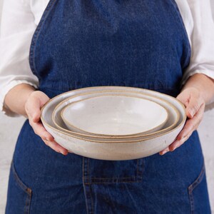 May include: A set of two nested ceramic bowls with a speckled off-white glaze and exposed brown clay rims. The bowls have a shallow, wide design with smooth, rounded edges, showcasing a natural, earthy texture on the exterior and interior surfaces.