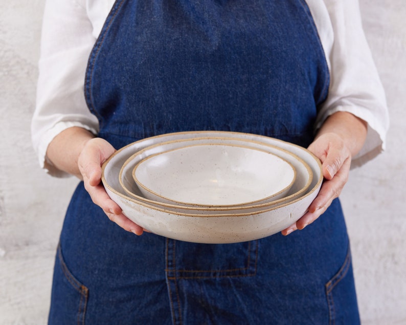 May include: A set of two nested ceramic bowls with a speckled off-white glaze and exposed brown clay rims. The bowls have a shallow, wide design with smooth, rounded edges, showcasing a natural, earthy texture on the exterior and interior surfaces.