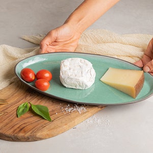 May include: A light blue oval ceramic serving platter with a dark gray rim, holding a round white cheese, a wedge of yellow cheese, and three red cherry tomatoes.