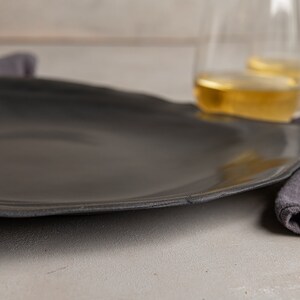 May include: A large, matte black dinner plate with an organic, slightly irregular circular shape and a smooth surface. The plate features a subtle, textured rim that follows the natural contour of the edge, providing a simple and functional design.