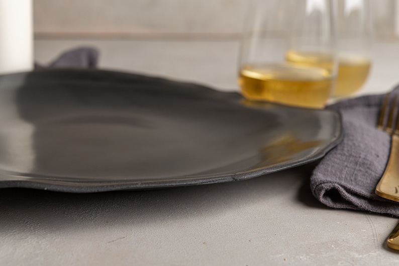 May include: A large, matte black dinner plate with an organic, slightly irregular circular shape and a smooth surface. The plate features a subtle, textured rim that follows the natural contour of the edge, providing a simple and functional design.