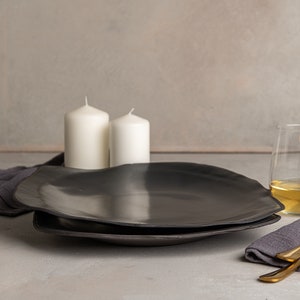 May include: Two matte black ceramic plates with irregular, organic edges stacked together. The setting includes two white pillar candles, two clear glass tumblers filled with a light-colored liquid, two gold-toned forks, and dark gray linen napkins.