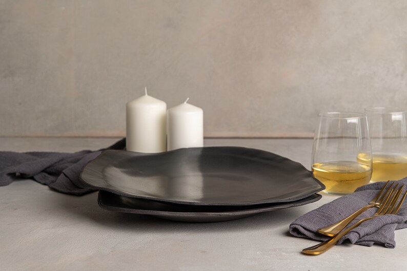 May include: Two matte black ceramic plates with irregular, organic edges stacked together. The setting includes two white pillar candles, two clear glass tumblers filled with a light-colored liquid, two gold-toned forks, and dark gray linen napkins.