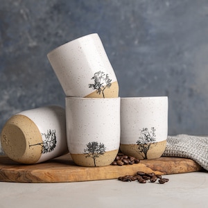 Peut inclure: Quatre tasses en céramique blanche avec un glaçage moucheté et un bord brun. Chaque tasse présente une silhouette noire d'un arbre. Les tasses sont empilées sur une planche à découper en bois avec des grains de café éparpillés autour.