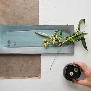 May include: A rectangular blue ceramic serving tray with a tree design and a bouquet of white flowers on top. A small black bowl with white flowers inside is held by a hand.