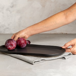 May include: Three red plums on a black oval plate, resting on a gray linen napkin. The plate is being placed on a gray surface.