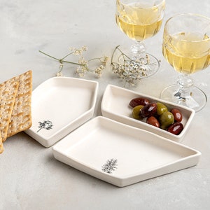 May include: Three white ceramic serving dishes with a simple tree design. The dishes are arranged on a light grey surface with a stack of crackers and olives in one of the dishes. Two glasses of white wine are in the background.