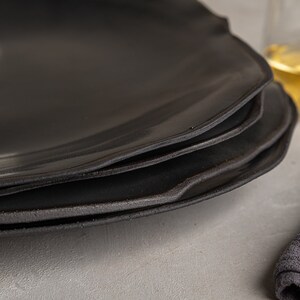 May include: A stack of three matte black plates with irregular, organic edges. The plates feature a smooth, dark surface with a slightly textured, raw-looking rim. The design is simple and functional, showcasing a dark, monochromatic aesthetic.