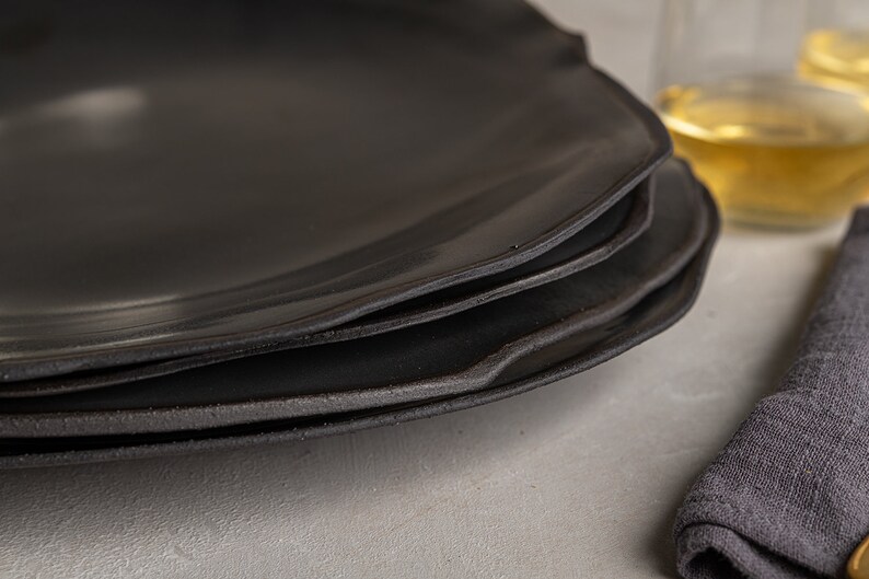 May include: A stack of three matte black plates with irregular, organic edges. The plates feature a smooth, dark surface with a slightly textured, raw-looking rim. The design is simple and functional, showcasing a dark, monochromatic aesthetic.