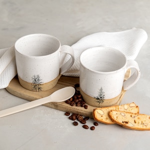 Puede incluir: Dos tazas de cerámica blanca con un diseño de árbol y un borde marrón, sentadas sobre una tabla de madera con granos de café y galletas biscotti.