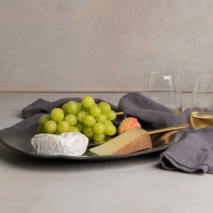 May include: A dark gray ceramic platter holds a cluster of green grapes, a wedge of firm cheese with a textured rind, a round soft cheese, and a lychee. A dark gray linen napkin, two glasses of white wine, and two white pillar candles are nearby.