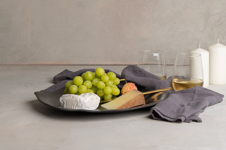 May include: A dark gray ceramic platter holds a cluster of green grapes, a wedge of firm cheese with a textured rind, a round soft cheese, and a lychee. A dark gray linen napkin, two glasses of white wine, and two white pillar candles are nearby.