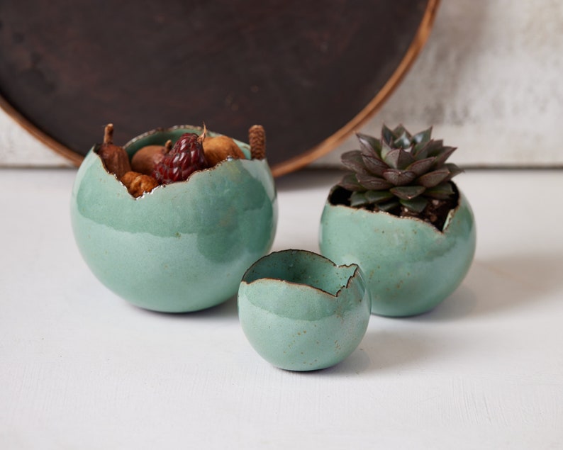 May include: Three teal green ceramic bowls with a crackled finish. The largest bowl is filled with nuts and dried fruit. The smallest bowl is empty. The middle bowl has a small succulent plant inside.
