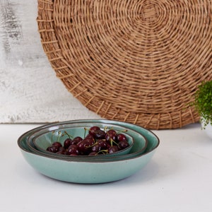 May include: Two nested round ceramic bowls with a smooth light teal glaze and dark brown rims. The bowls are filled with a cluster of fresh dark red cherries with green stems, showcasing a simple design and a speckled texture on the exterior.