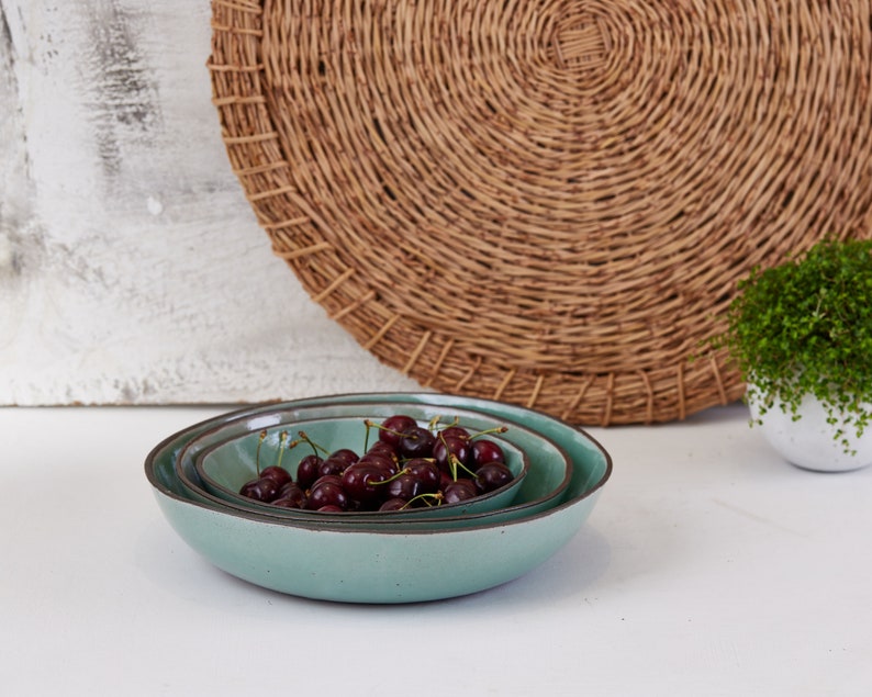 May include: Two nested round ceramic bowls with a smooth light teal glaze and dark brown rims. The bowls are filled with a cluster of fresh dark red cherries with green stems, showcasing a simple design and a speckled texture on the exterior.