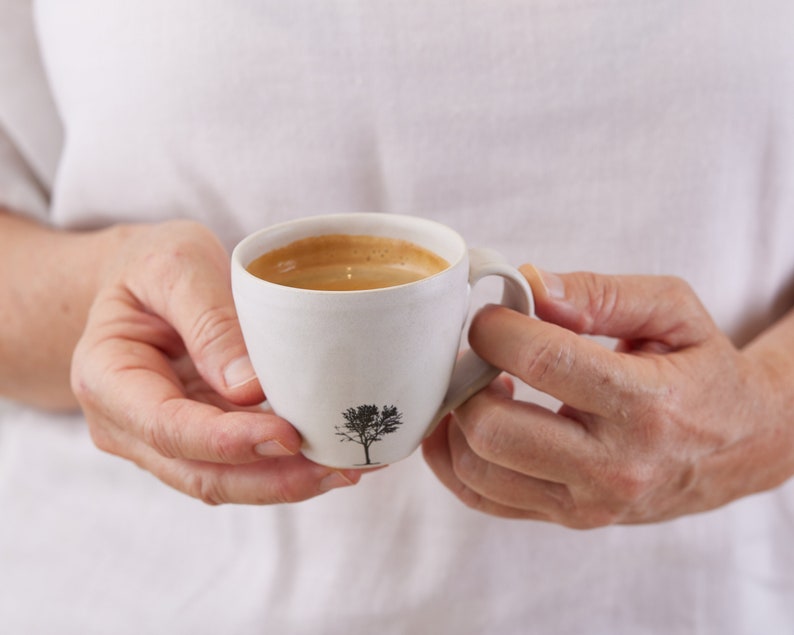 以下が含まれることがあります： 表面に質感のあるマットなオフホワイトの陶器製コーヒーカップで、前面に小さな黒い木のシルエットがプリントされています。カップには表面に薄茶色のクレマ層があるダークブラウンのコーヒーが入っており、持ち手が付いています。