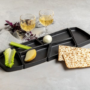 May include: A black divided serving platter with a hard-boiled egg, potato, celery, lettuce, and two pieces of matzah. Two glasses of white wine are on the table next to the platter.