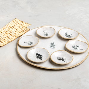May include: A white ceramic Seder plate with six small bowls, each featuring a different tree design. The plate has a speckled glaze and a brown rim. A piece of matzah is in the background.