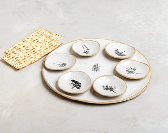 May include: A white ceramic Seder plate with a speckled finish and a tan rim, featuring six small, shallow, circular indentations, each decorated with a unique black botanical illustration, and a central tree design, accompanied by two pieces of matzah.