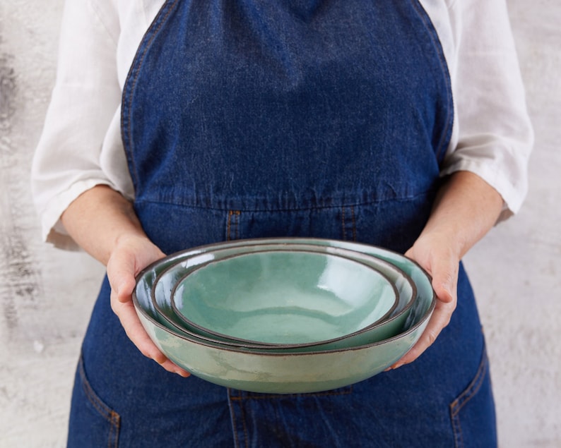 May include: A set of two nested ceramic bowls with a smooth, light seafoam green glaze and a dark, contrasting rim. The bowls feature a rounded, organic shape with a glossy finish, stacked one inside the other to display their varying sizes.