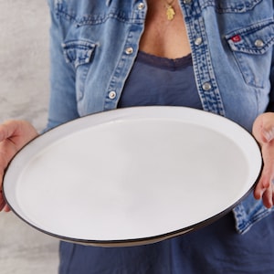 Peut inclure: Un grand plat de service rond blanc avec un bord fin brun foncé. Le plat présente une finition lisse et brillante ainsi qu'une surface plate et peu profonde, conçue pour présenter des aliments. Les bords sont légèrement surélevés.