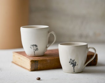 May include: Two off-white ceramic mugs with a smooth, slightly irregular finish. Each mug features a small, detailed black silhouette of a tree printed near the base. One mug rests on a vintage brown hardcover book, while the other sits beside it.