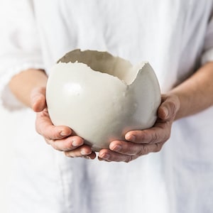 Może przedstawiać: Biała ceramiczna doniczka z dużą szczeliną po jednej stronie. Doniczka ma kształt kuli i jest trzymana w czyichś rękach.