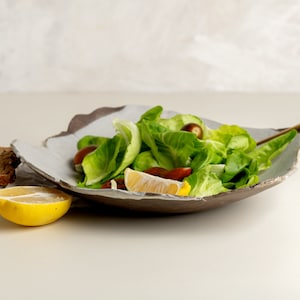 Könnte beinhalten: Eine blattförmige Keramikschale gefüllt mit frischem Salat, Kirschtomaten und einem Zitronenkeil. Ein goldener Löffel liegt in der Schale. Brotscheiben und eine Gabel liegen daneben, was auf eine gesunde Mahlzeit hindeutet.