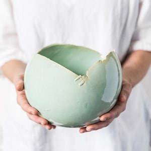 May include: A light green ceramic bowl with a crack on the side. The bowl is round and has a smooth, glossy finish.